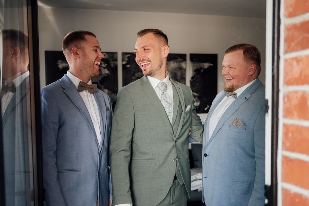 Groomsmen share laughter and conversation in formal suits while standing in a brick-walled hallway.