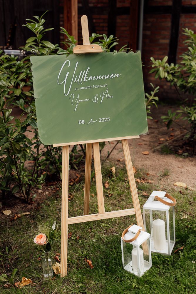 Wooden easel displays elegant green welcome sign with white lettering next to decorative lanterns outdoors.