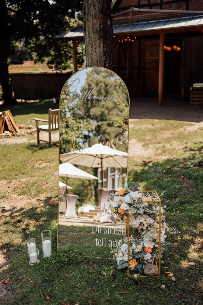 A decorative arched mirror with welcome text stands outdoors by a rustic barn, adorned with floral arrangements and candles.