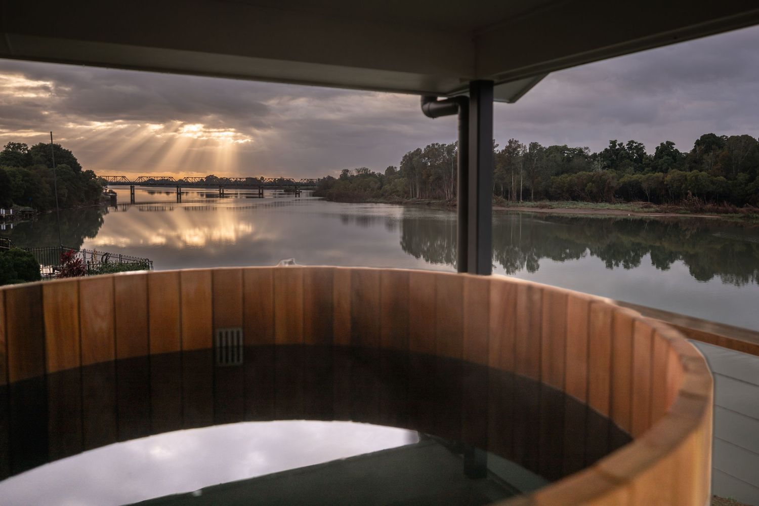 A wood-paneled hot tub overlooks a serene lake surrounded by trees at sunset with dramatic cloudy skies.