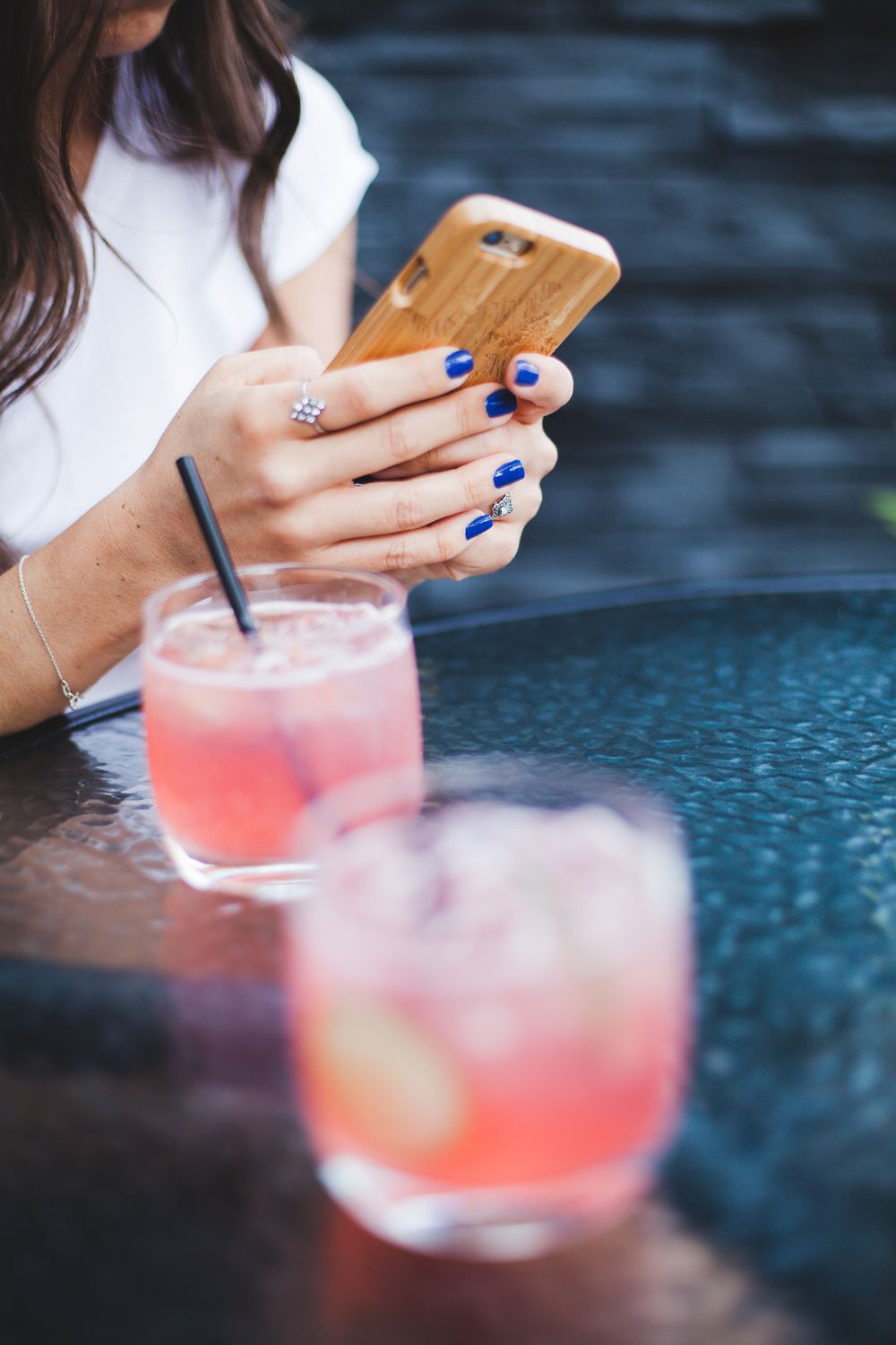Woman with blue nails using bamboo wood iPhone case at outdoor table with pink cocktails.