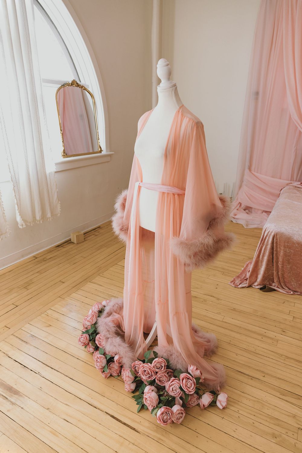 Pink feather-trimmed sheer robe on mannequin surrounded by roses in elegant studio.