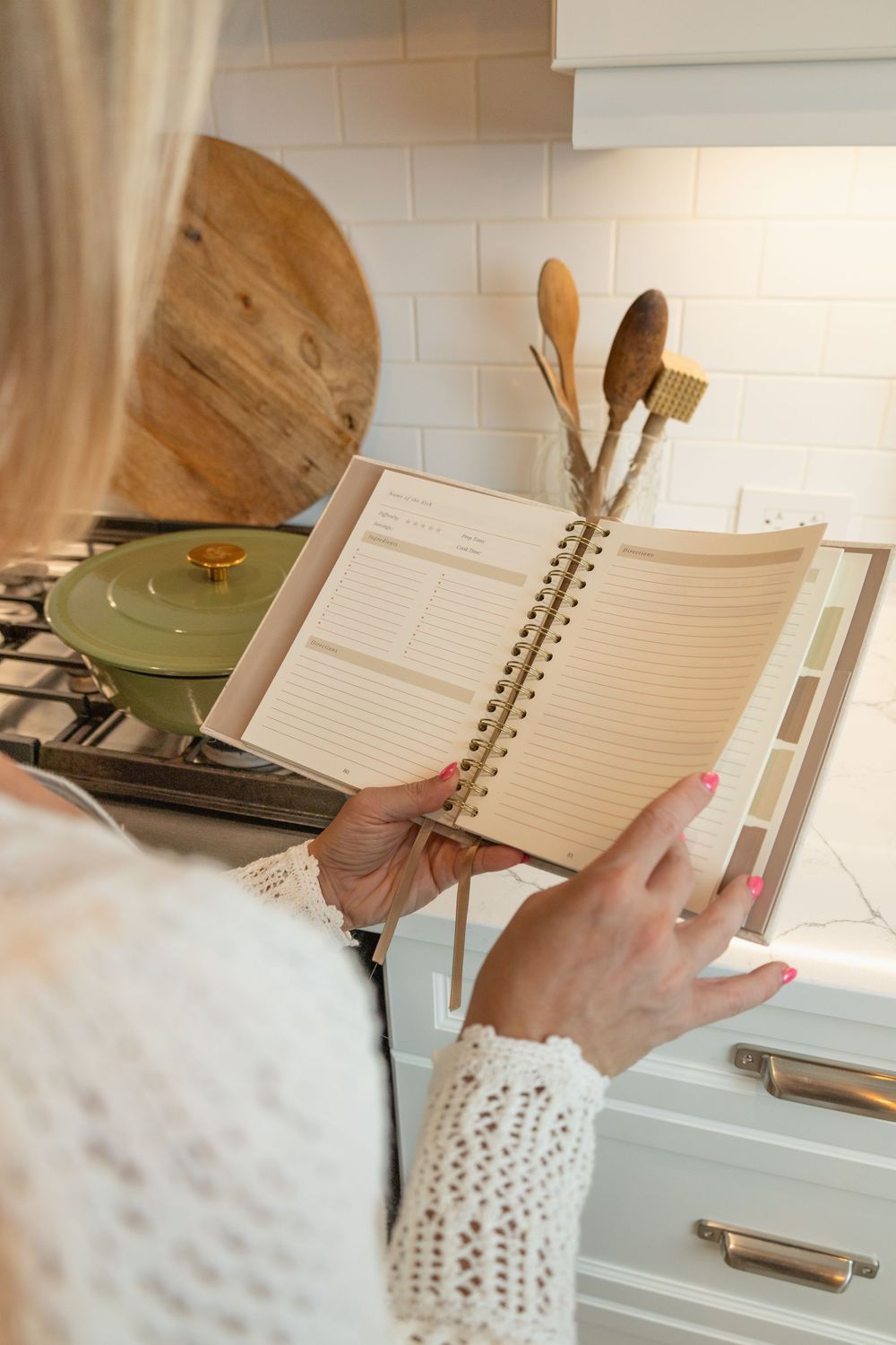 Blonde woman holding open a spiral-bound recipe journal near a gas stove in white kitchen.