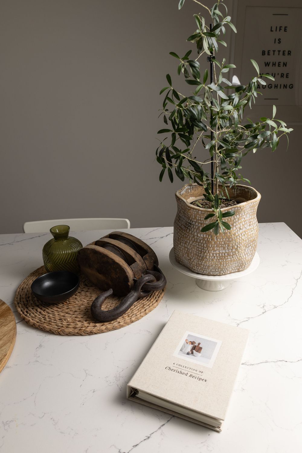 Cherished Recipes linen book on marble table beside potted olive tree and rustic wooden decor.