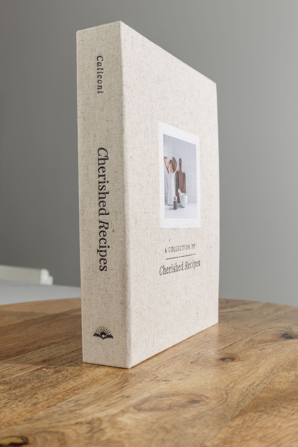Caliconi Cherished Recipes linen hardcover book standing upright on a wooden surface.
