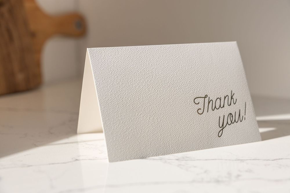 Elegant white embossed thank you card displayed on a marble surface in a bright kitchen setting.