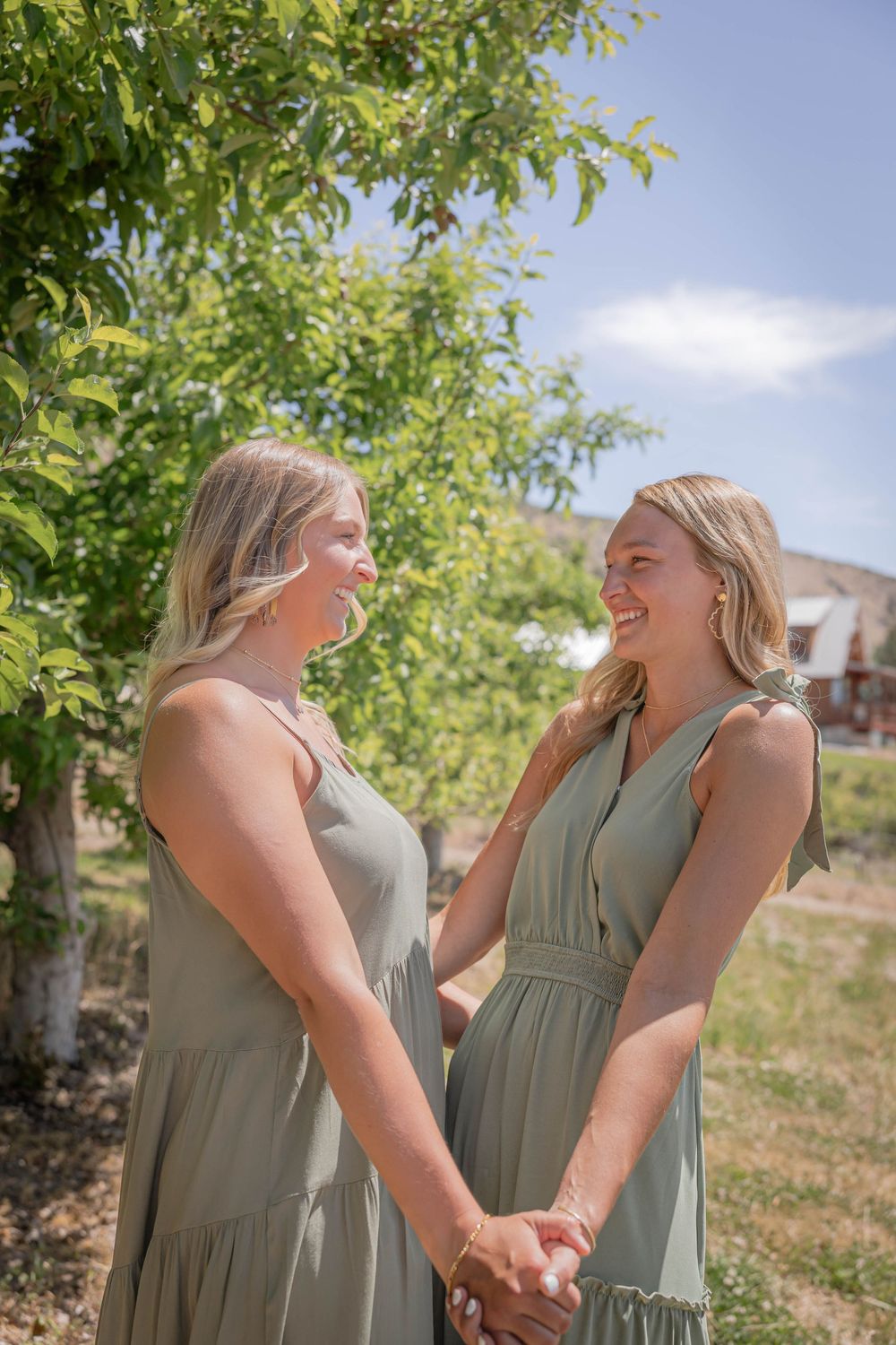 Two people in sage green dresses sharing a joyful moment outdoors under trees.