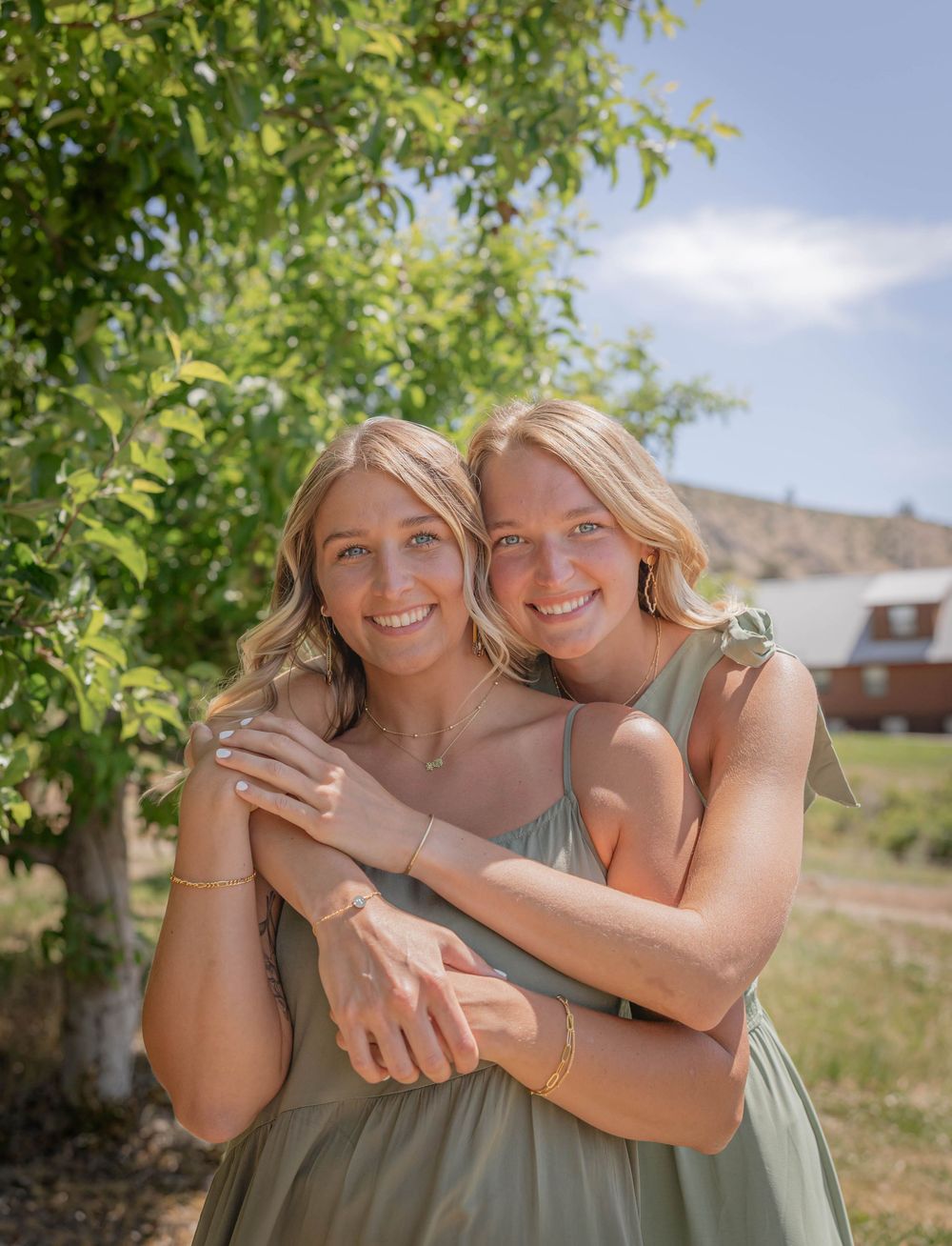 Two people in matching sage dresses embracing and laughing together under a tree.