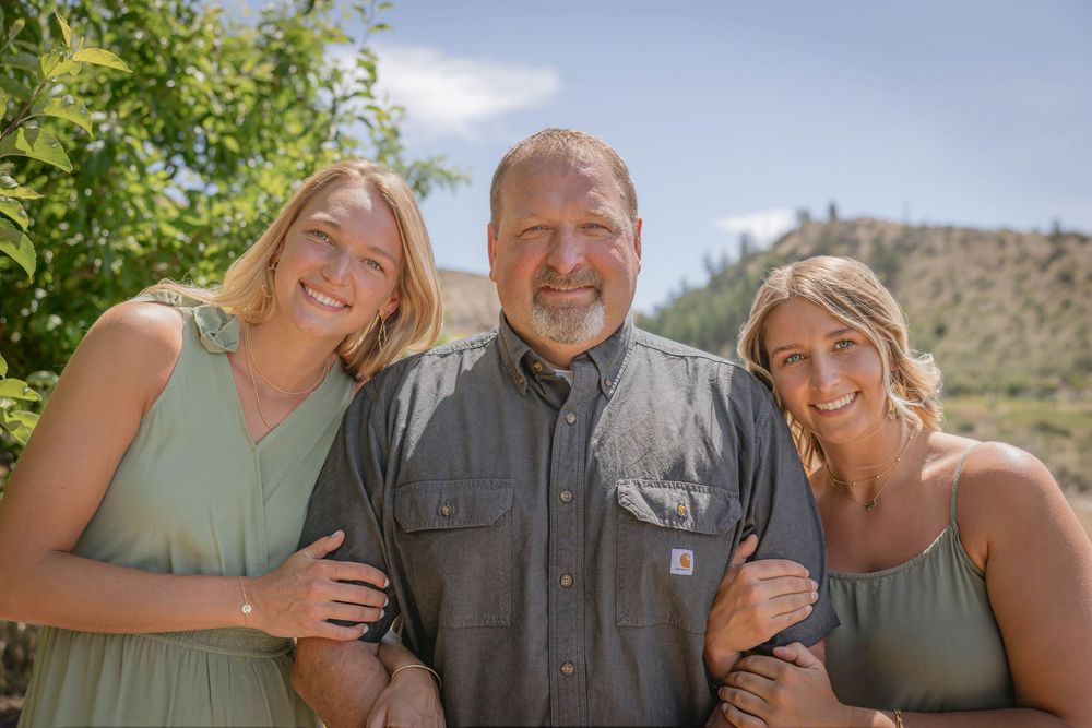 Three people smiling together outdoors with mountain scenery and trees in background.
