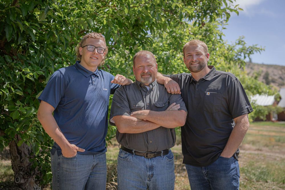 Three people stand close together for a photo under green trees on a bright summer day.