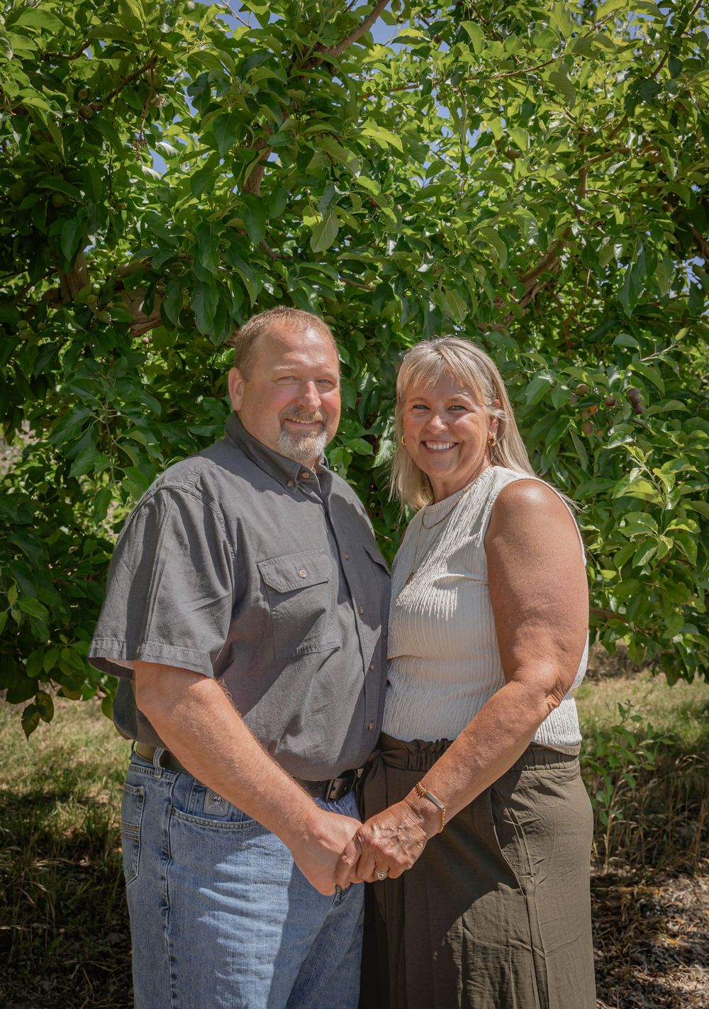 A couple embraces while posing for a photo outdoors with green foliage in the background.