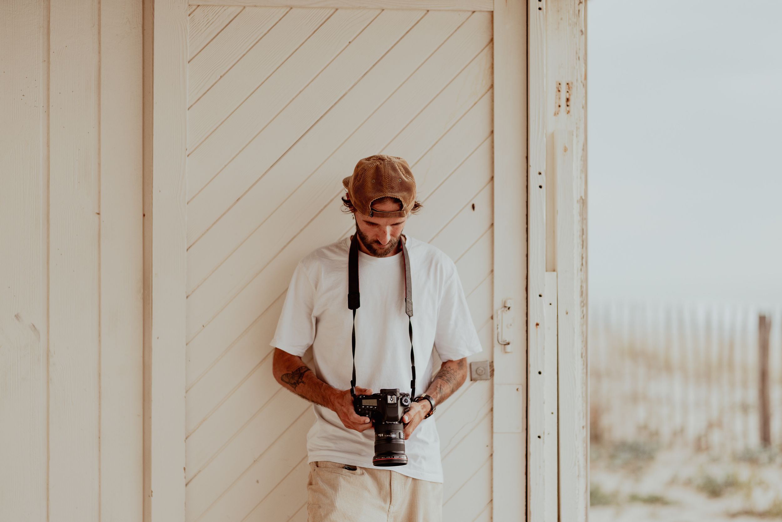 Photographer in white attire holding camera while standing against wooden beach wall.