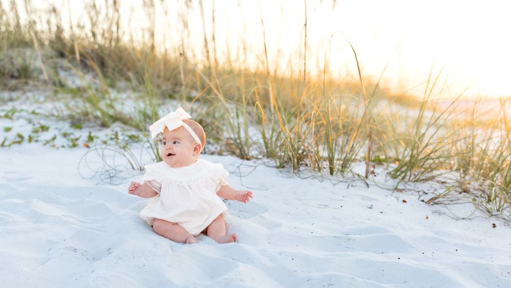 Baby in white dress sits on sandy beach at sunset while playing with sand and sea grass.