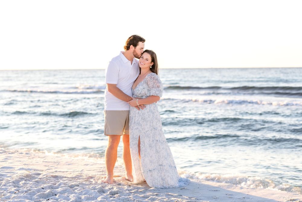 A couple embraces while standing barefoot on a white sandy beach at sunset with ocean waves in the background.