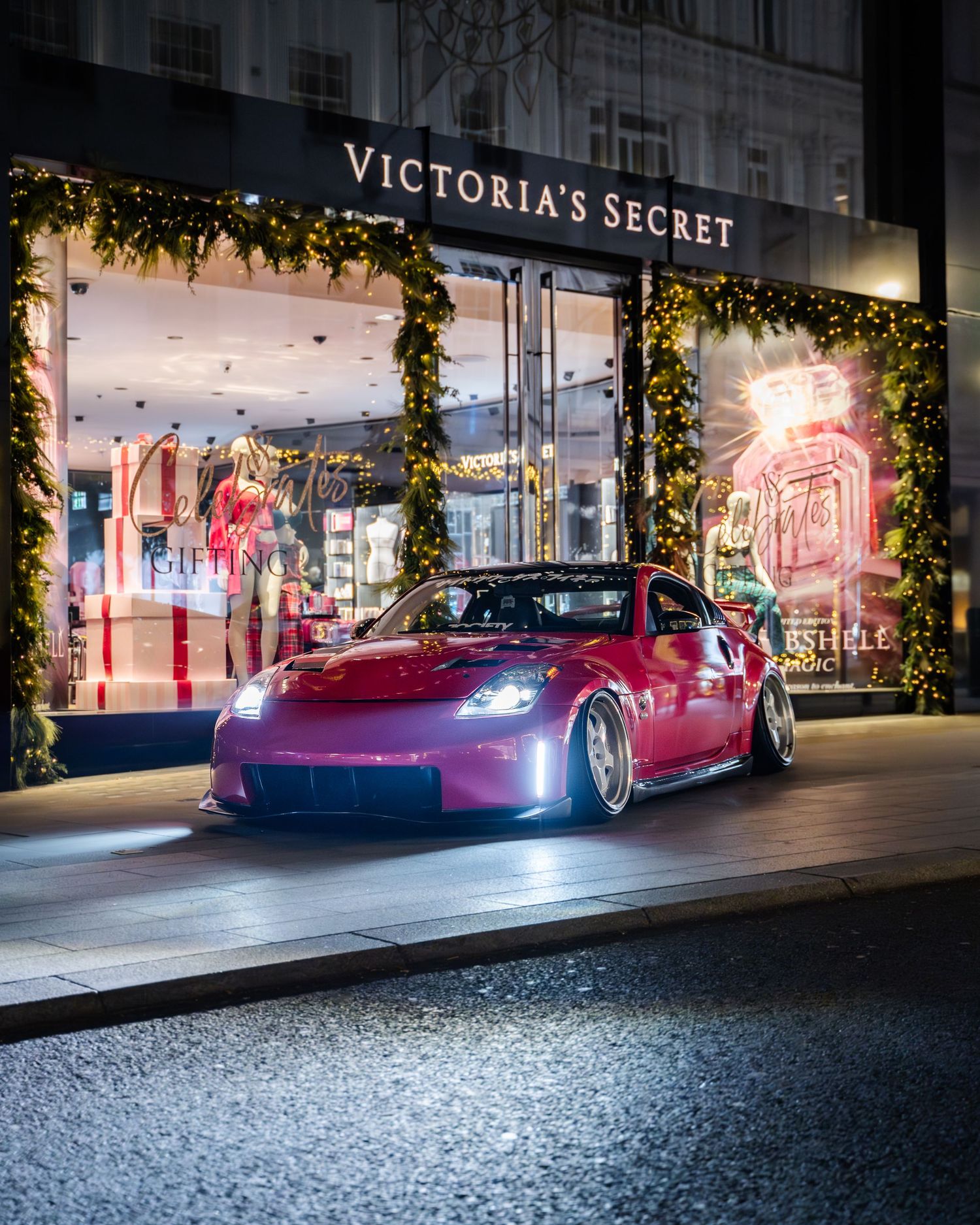 Pink sports car parked outside Victoria's Secret store at night with illuminated storefront.