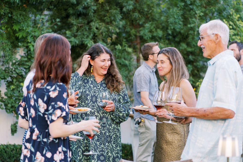 Group of people enjoying drinks and conversation at an outdoor garden party with lush greenery in the background.