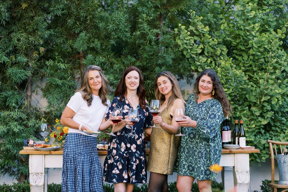 A group of friends posing together outdoors in front of green foliage wearing casual summer attire.