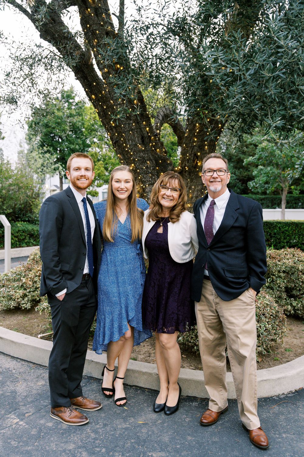 A group of people in formal attire stand together outdoors near trees and a white railing.