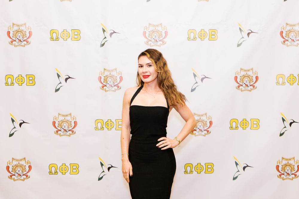 Woman in black dress poses on red carpet with gold and white step-and-repeat backdrop.