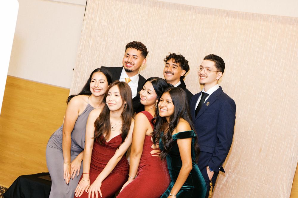A group of friends in formal attire pose together at a special event against a beige and yellow wall.