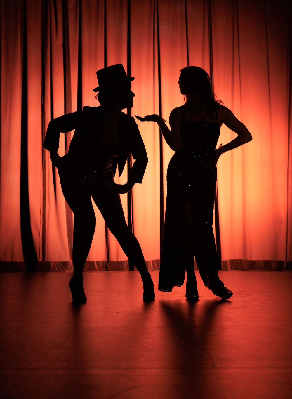 Silhouette of two dancers performing on a warmly lit stage with orange curtain backdrop.