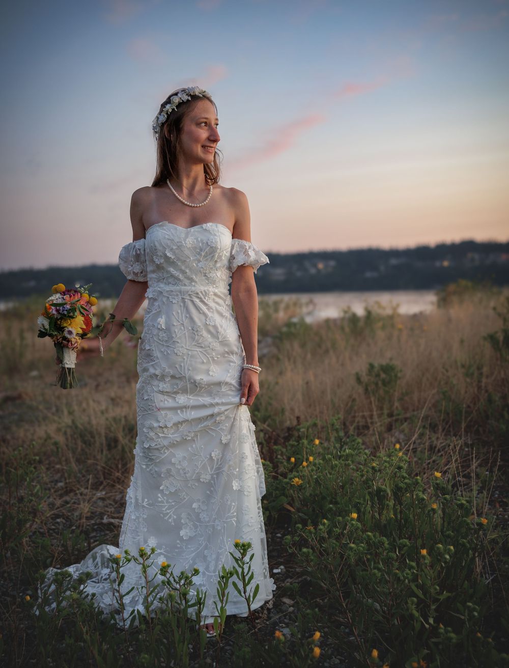 Bride in off-shoulder lace wedding gown holding colorful bouquet at sunset near a scenic waterfront.
