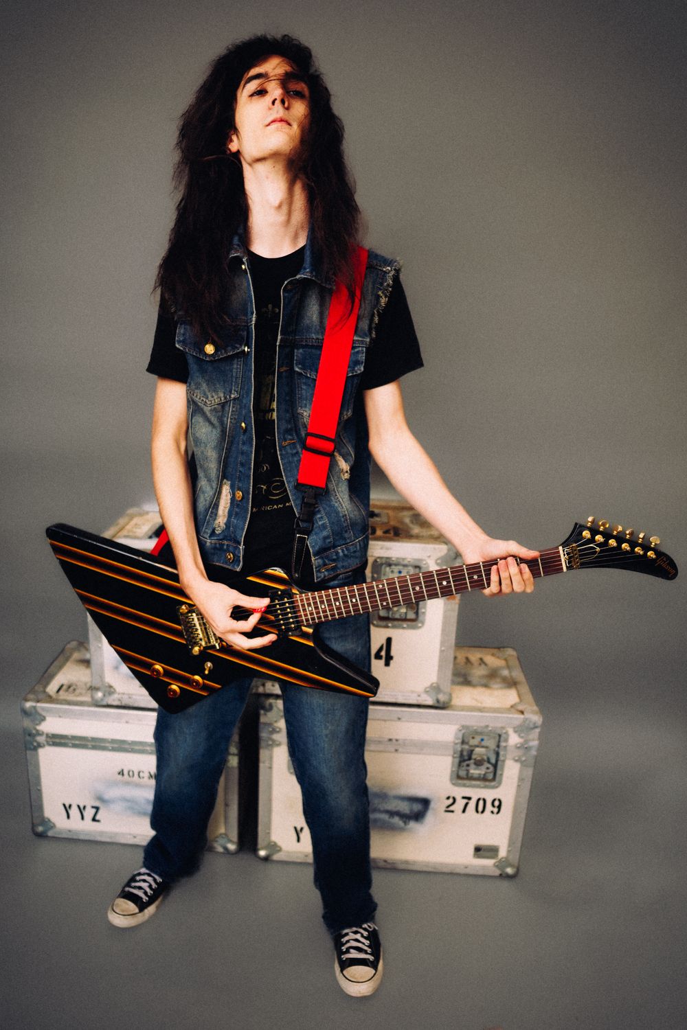 Young rock guitarist with long hair playing electric guitar in denim vest against gray background.