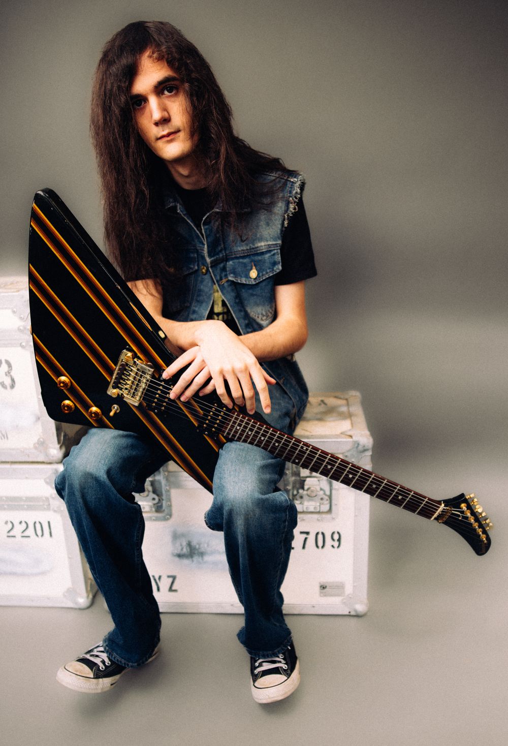 Young male guitarist with long hair sitting on road cases holding a black and gold electric guitar.