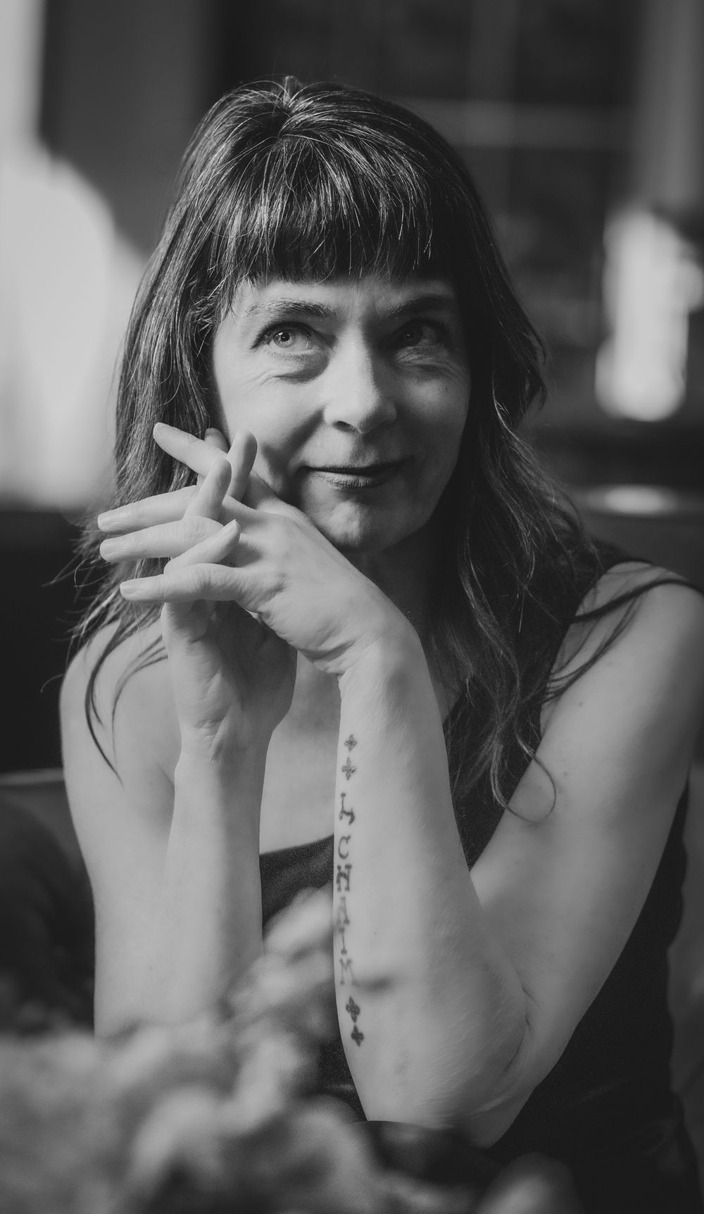 Black and white portrait of a woman with bangs and forearm tattoo resting her chin on her hands.