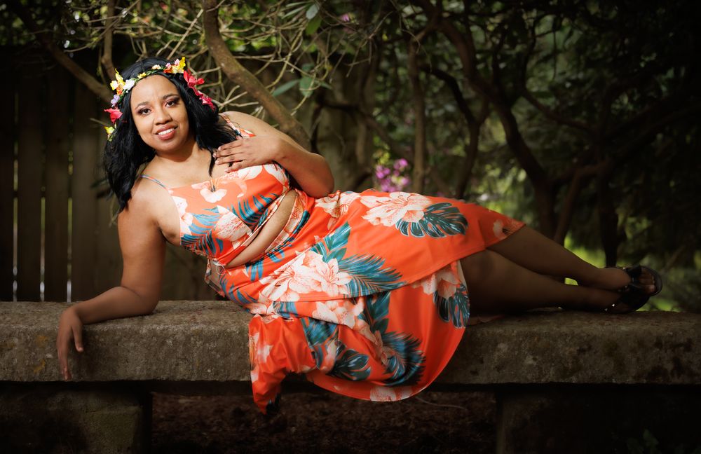 Woman in orange floral strapless dress with flower crown reclining gracefully on a stone wall outdoors.