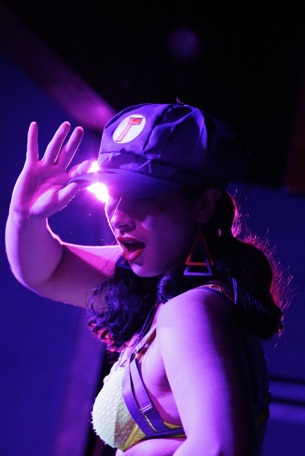 Female performer on stage under purple lights wearing a cap and raising her hand dramatically.