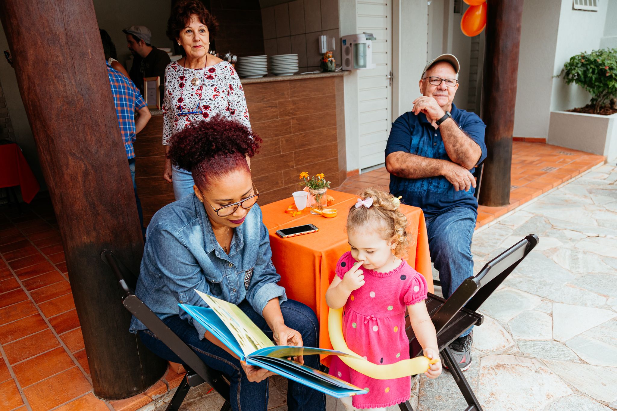 Adult reading a storybook with small child at outdoor restaurant while others sit at orange table nearby.