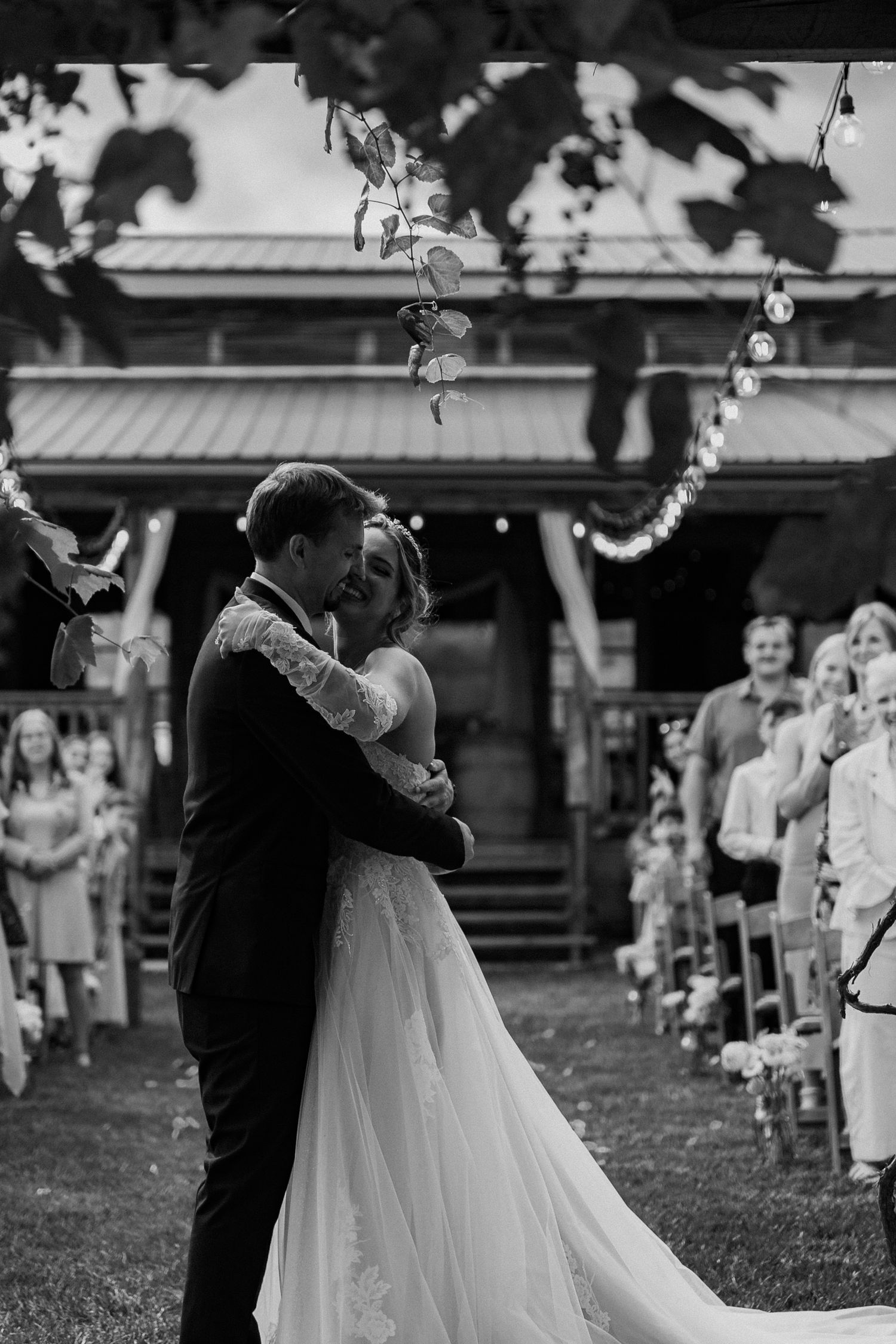 A couple shares their first dance at an outdoor wedding ceremony with guests watching and string lights overhead.