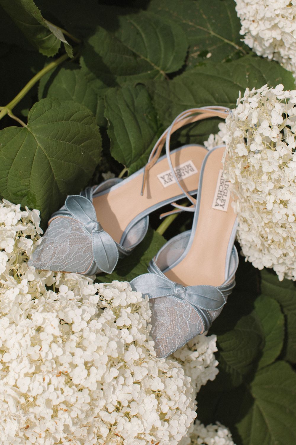 Elegant silver butterfly wedding shoes displayed among white hydrangea flowers and green leaves.