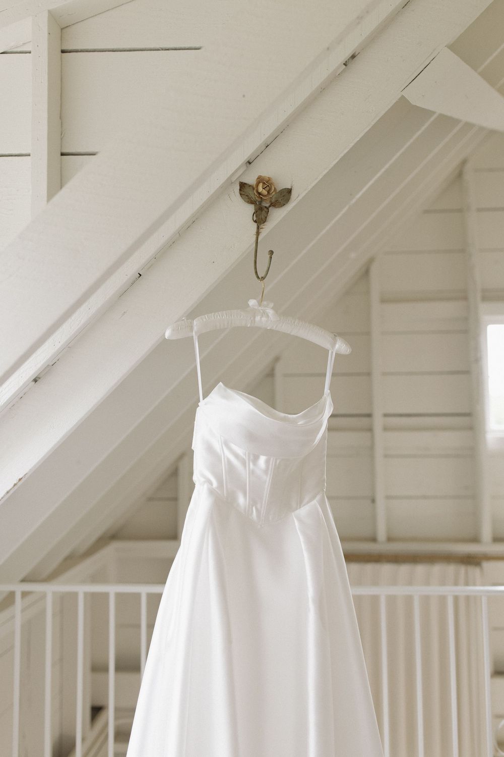 White wedding dress hanging from wooden beams in a bright white room with sloped ceiling.