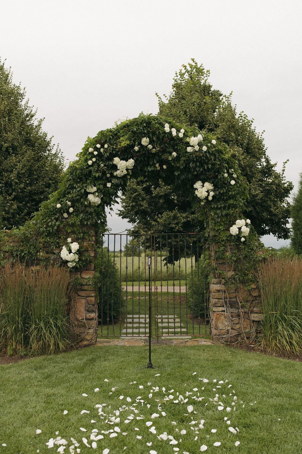 Stone garden entrance with white floral arch and scattered rose petals creating romantic wedding setting.