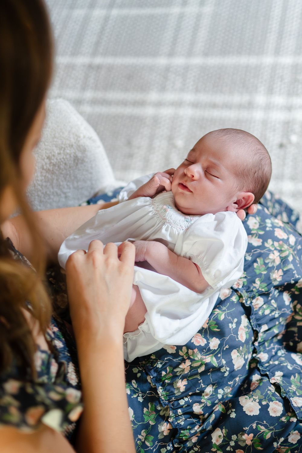 A sleeping newborn baby wearing white is cradled on a blue floral blanket while being fed with a bottle.