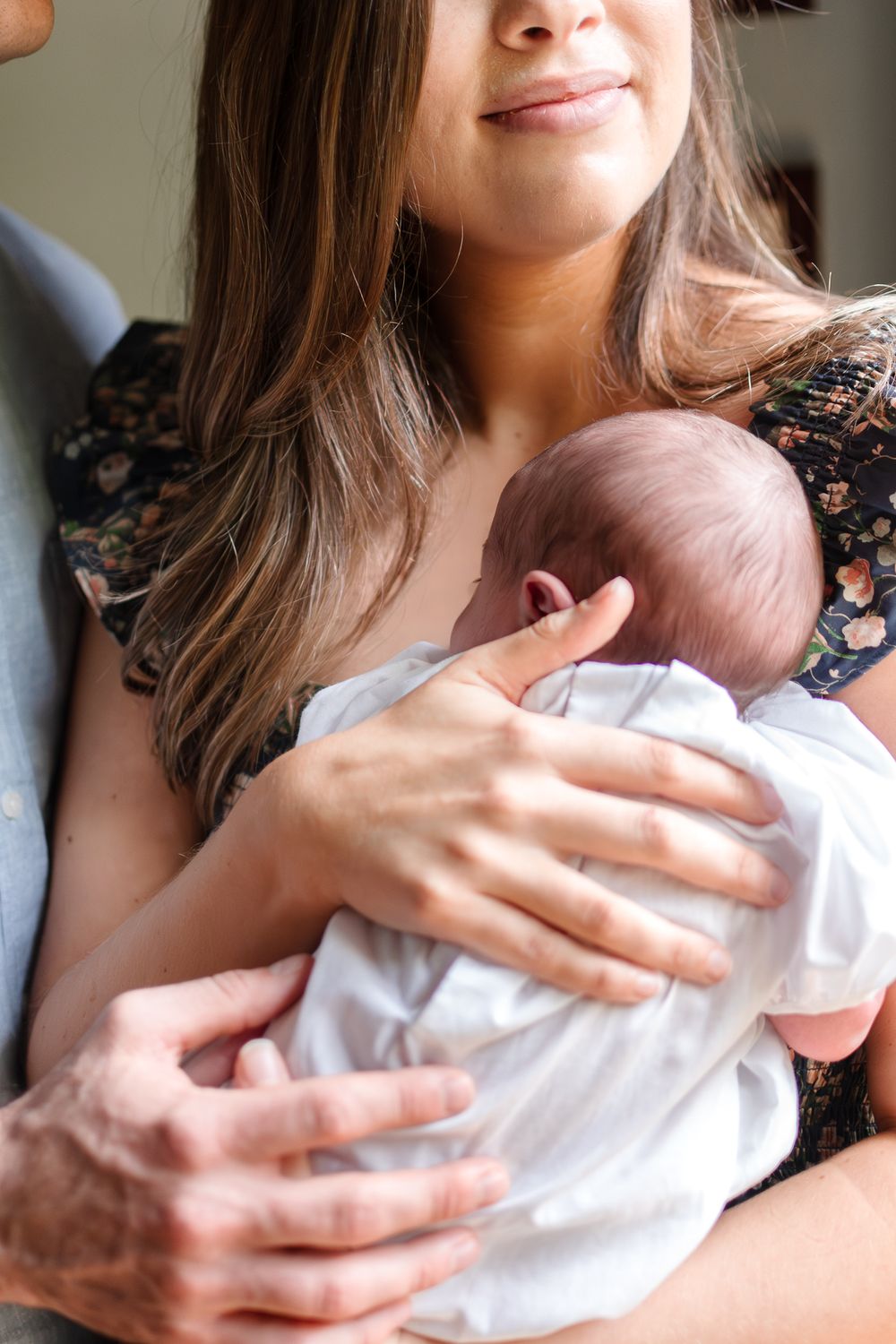 A tender moment captured as caring hands hold a newborn baby wrapped in white against a floral print dress.