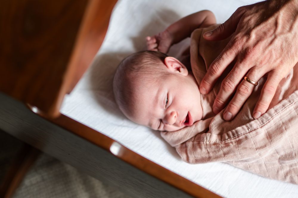 A sleeping newborn baby wrapped in peachy pink swaddle blanket lies peacefully on white bedding.