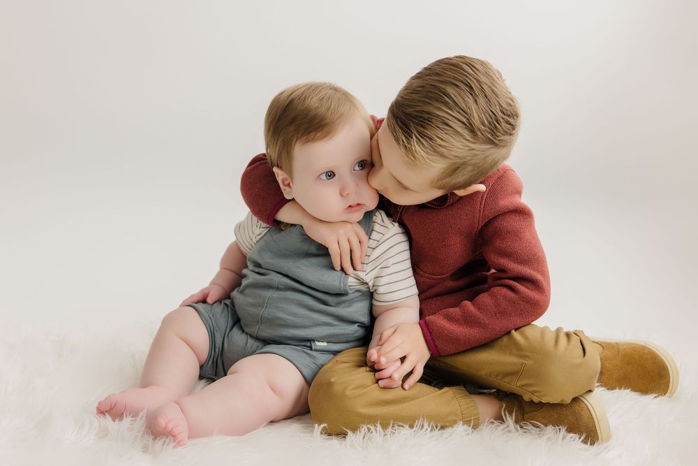 Two small children sitting on white carpet wearing burgundy and neutral colored clothing share a sweet moment together.