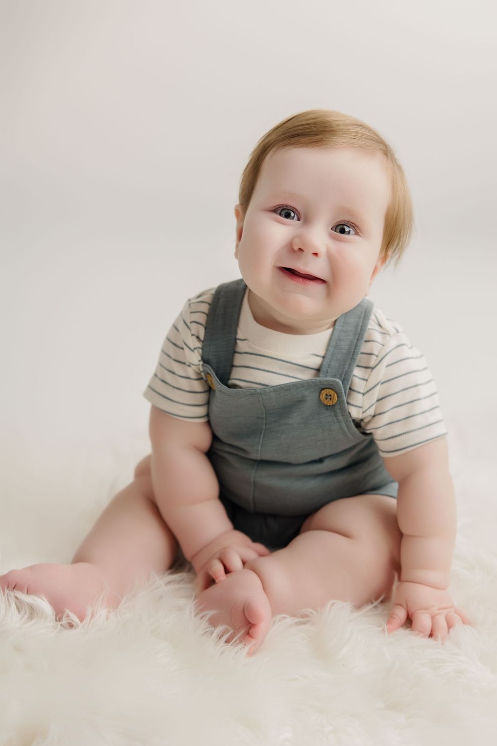 Baby in striped shirt and gray overalls smiling while sitting on a white fluffy rug.
