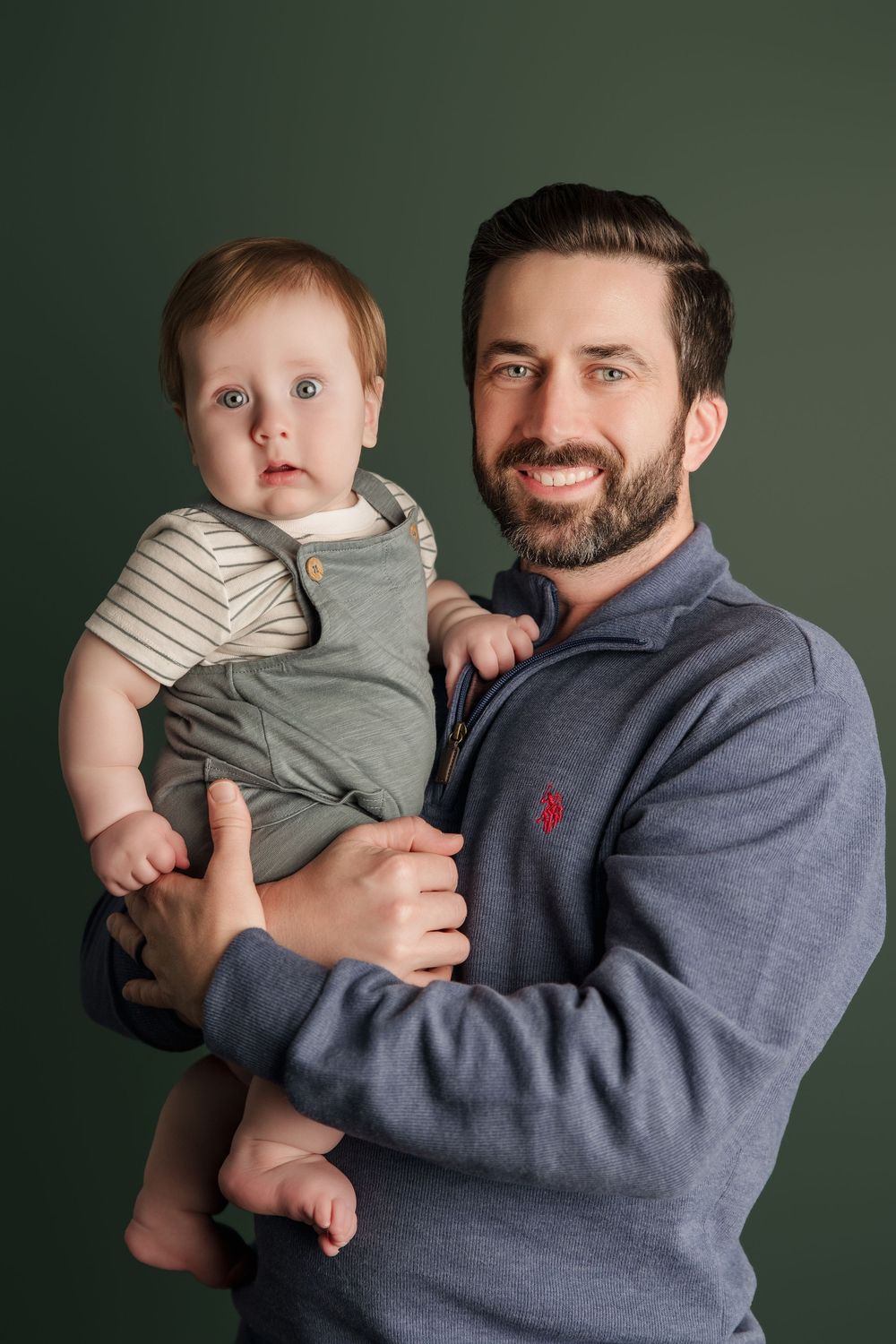 Portrait of a parent holding a baby wearing green overalls against a dark green background.
