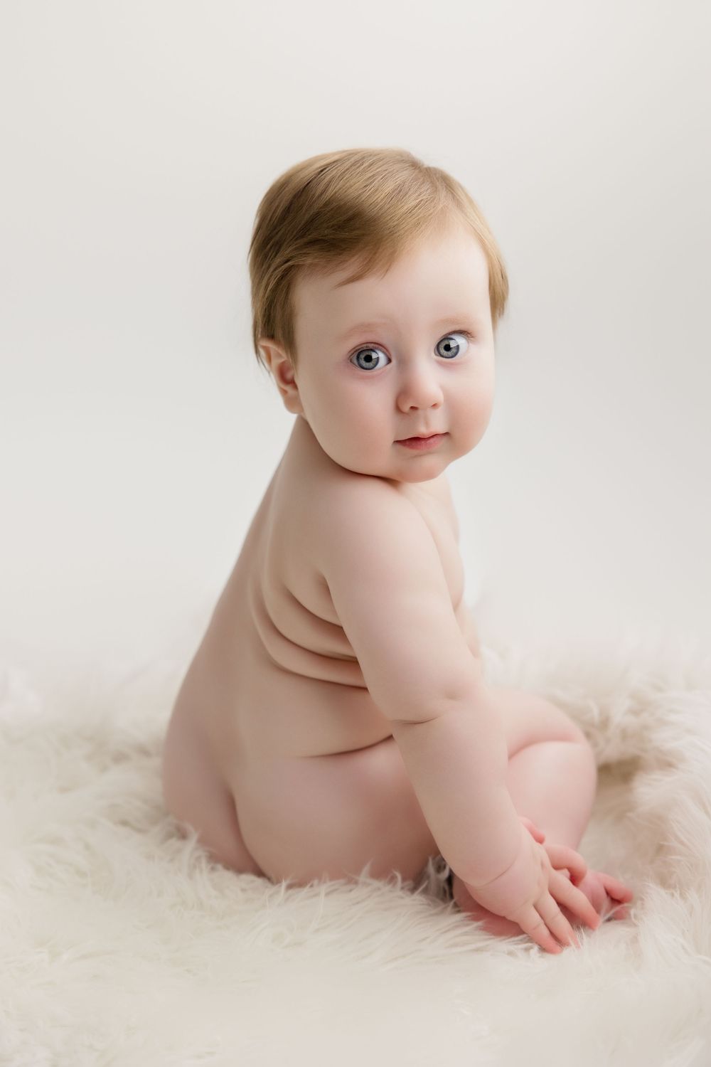 Adorable baby rolls over on soft white fur blanket in series of developmental milestone photos.