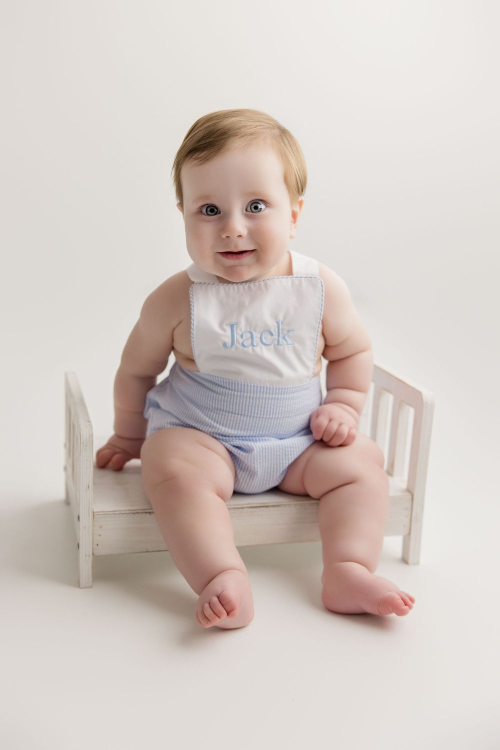 Baby wearing a light blue romper sits on a small white wooden bench.