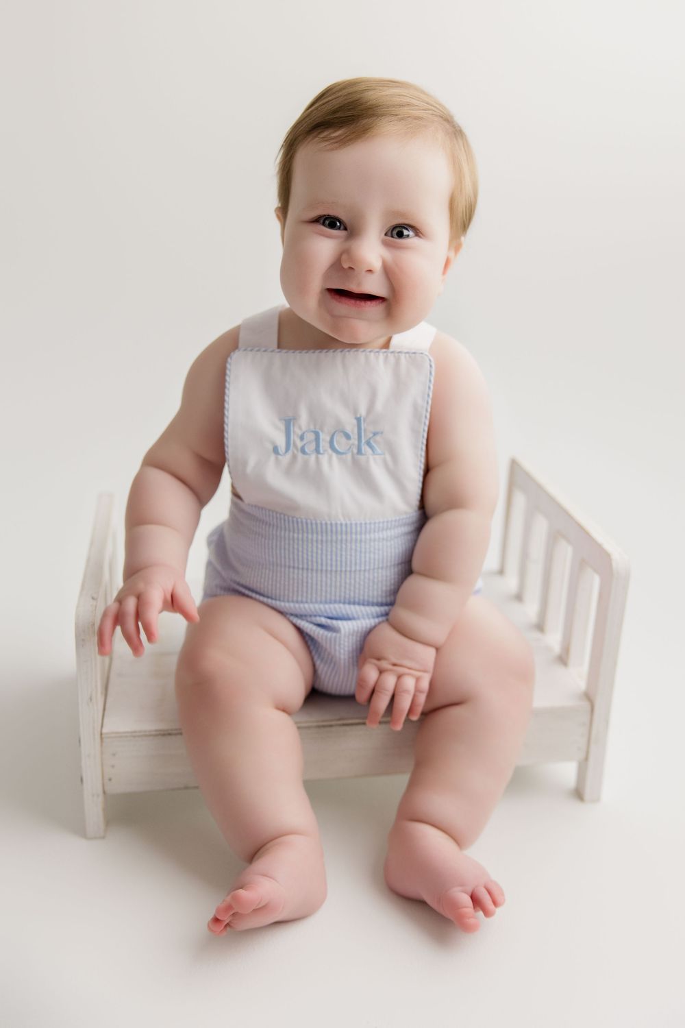 Baby in a white and blue sleeveless outfit sits smiling on a white wooden bench.