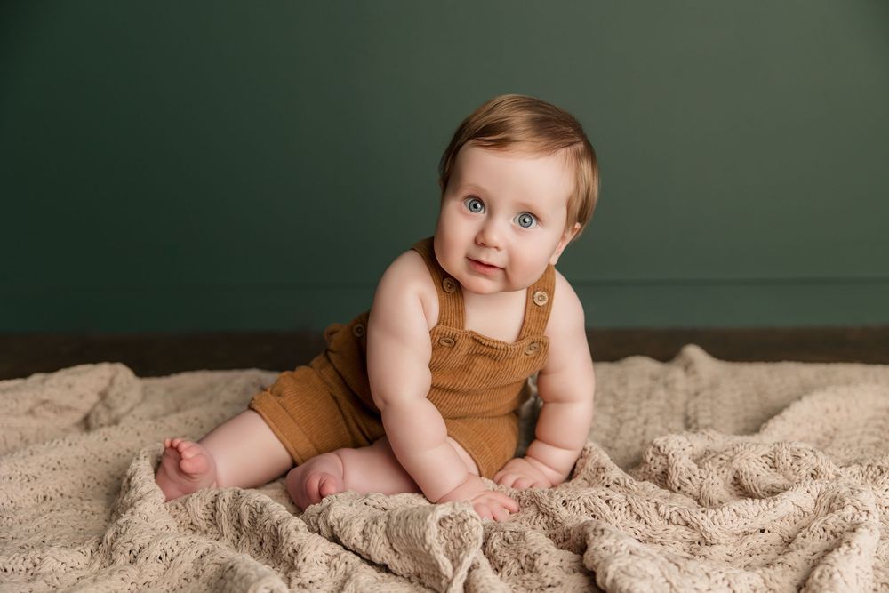 Baby in brown corduroy overalls sits on textured beige blanket against dark green wall background.