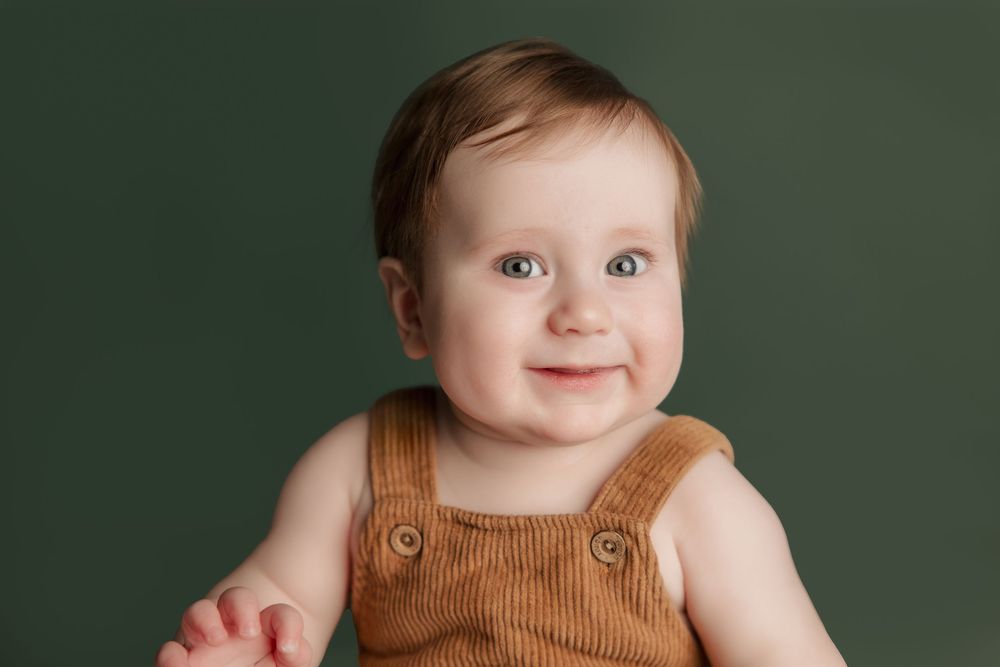 Baby wearing brown overalls sits against green background in multiple poses displaying various facial expressions.