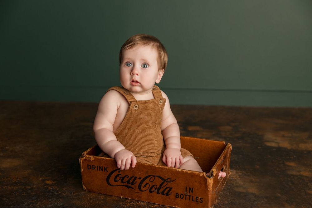 Baby sitting in vintage wooden Coca-Cola crates with rustic brown backdrop showing different poses.