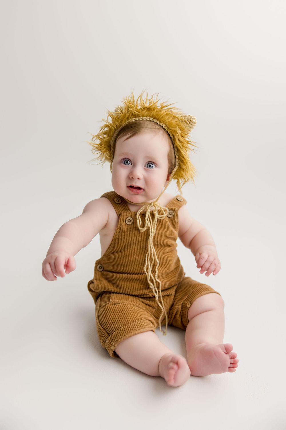 Baby in tan corduroy overalls and yellow fringe headpiece sits on white background.