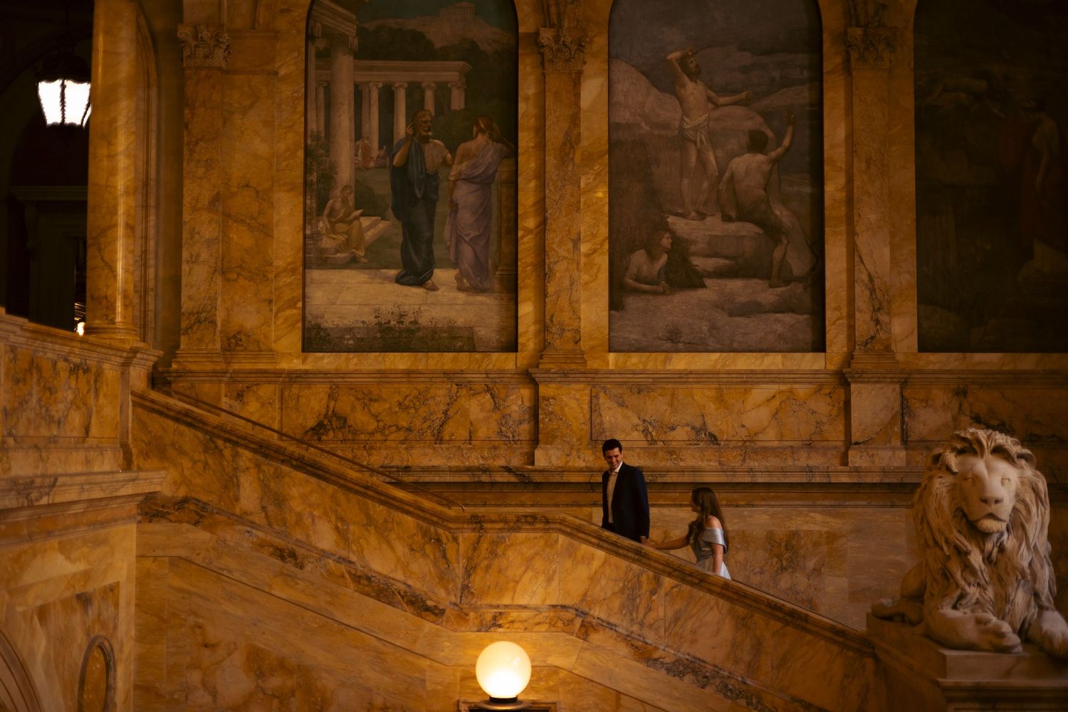 Two figures walk down an ornate marble staircase beneath classical wall paintings in a dimly lit historic building.
